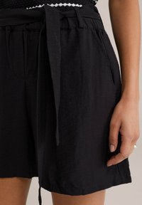 Shorts noirs en tissu léger, dotés d'une taille à nouer, de poches latérales et d'une coupe décontractée. La texture semble douce et légèrement froissée.