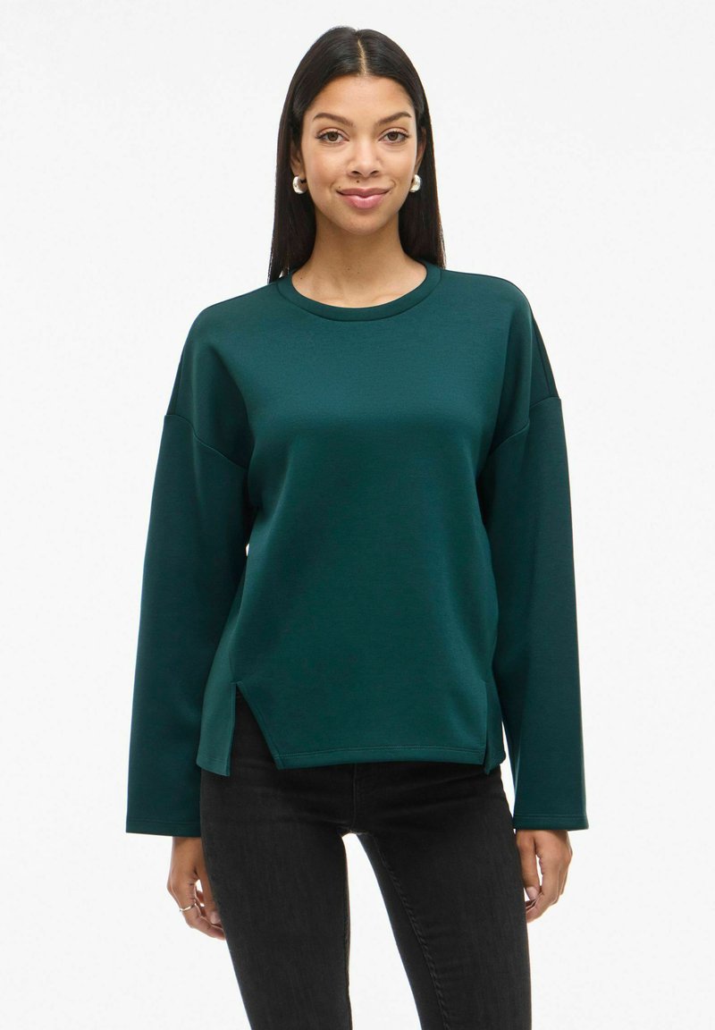 Sudadera de manga larga verde oscuro con cuello redondo, hombros caídos y aberturas laterales; textura suave, corte relajado.
