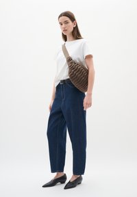 Borsa a tracolla marrone, con texture, indossata sopra una camicia bianca; abbinata a jeans a vita alta di colore blu scuro e scarpe nere a punta. Design semplice e moderno.