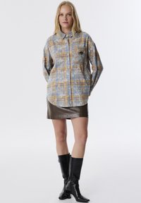 Camicia a maniche lunghe a quadri blu e gialli con chiusura a bottoni e taschino sul petto, abbinata a una minigonna in pelle marrone e stivali neri.