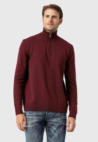 Burgunderfarbener Viertelreißverschluss-Pullover mit geribbeltem Kragen und Bündchen, aus strukturiertem Stoff gefertigt. Kombiniert mit blauen Jeans, die einen lässigen Look zeigen.