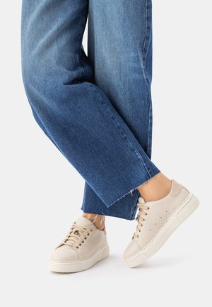 Stopy w kremowobiałych skórzanych trampkach z grubymi podeszwami i złotymi przelotkami, zestawione z przyciętymi niebieskimi dżinsami z postrzępionymi końcami.