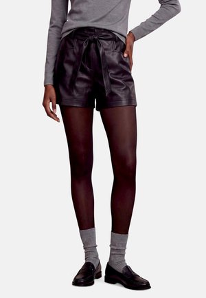 Mannequin portant un short en cuir noir à nouer à la taille, un haut gris et des chaussettes, des collants foncés et des mocassins noirs sur un fond uni.