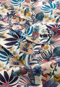 Poignet de chemise boutonné avec imprimé floral tropical coloré comprenant des ananas, des hibiscus, des feuilles et des bananes sur un tissu clair.