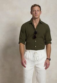 Camisa de botões verde-escura com mangas arregaçadas, combinada com umas calças brancas com cordão. Acessorizada com óculos de sol pretos e um relógio.