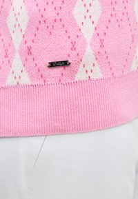 Πλεκτό πουλόβερ με ροζ και λευκό σχέδιο argyle, με μια μικρή μαύρη μεταλλική ετικέτα με την επιγραφή «Dalma» πάνω από λευκό παντελόνι.