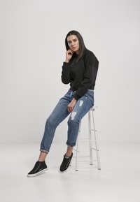 Felpe nere con maniche testurizzate, jeans blu strappati e sneaker nere. Modello seduto su uno sgabello bianco contro uno sfondo semplice.