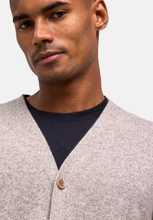 Cardigan beige chiaro in materiale lavorato a maglia con scollo a V, caratterizzato da una singola chiusura con bottone in legno e indossato sopra una camicia scura.