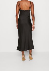 Robe en satin noir à fines bretelles, texture lisse, et silhouette évasée en ligne A. Associée à des sandales à talons hauts multicolores.