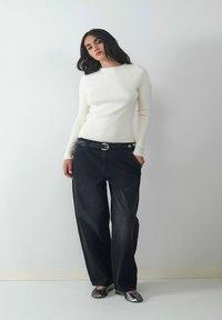 Kobieta z długimi ciemnymi włosami, ubrana w biały sweter z prążkowanym wzorem, luźne czarne spodnie z paskiem i metaliczne srebrne baleriny, stojąca przy gładkiej ścianie.