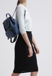 Femme portant un cardigan court blanc et une jupe noire, portant un sac à dos en cuir bleu marine sur l'épaule.