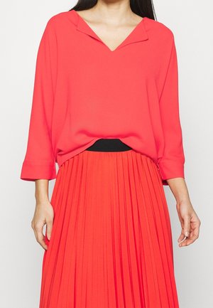 Woman wearing a loose red V-neck blouse with three-quarter sleeves and a high-waisted red pleated skirt with a black waistband.