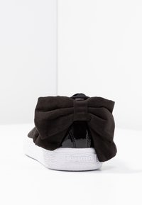 Sneaker in suede nera con un grande dettaglio a fiocco sul tallone, accenti neri lucidi e una suola in gomma bianca testurizzata.