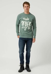 Hombre de pie que lleva un sudadera verde con un gráfico de ancla y gorra de marinero, junto con jeans azul marino y botas negras con cordones.