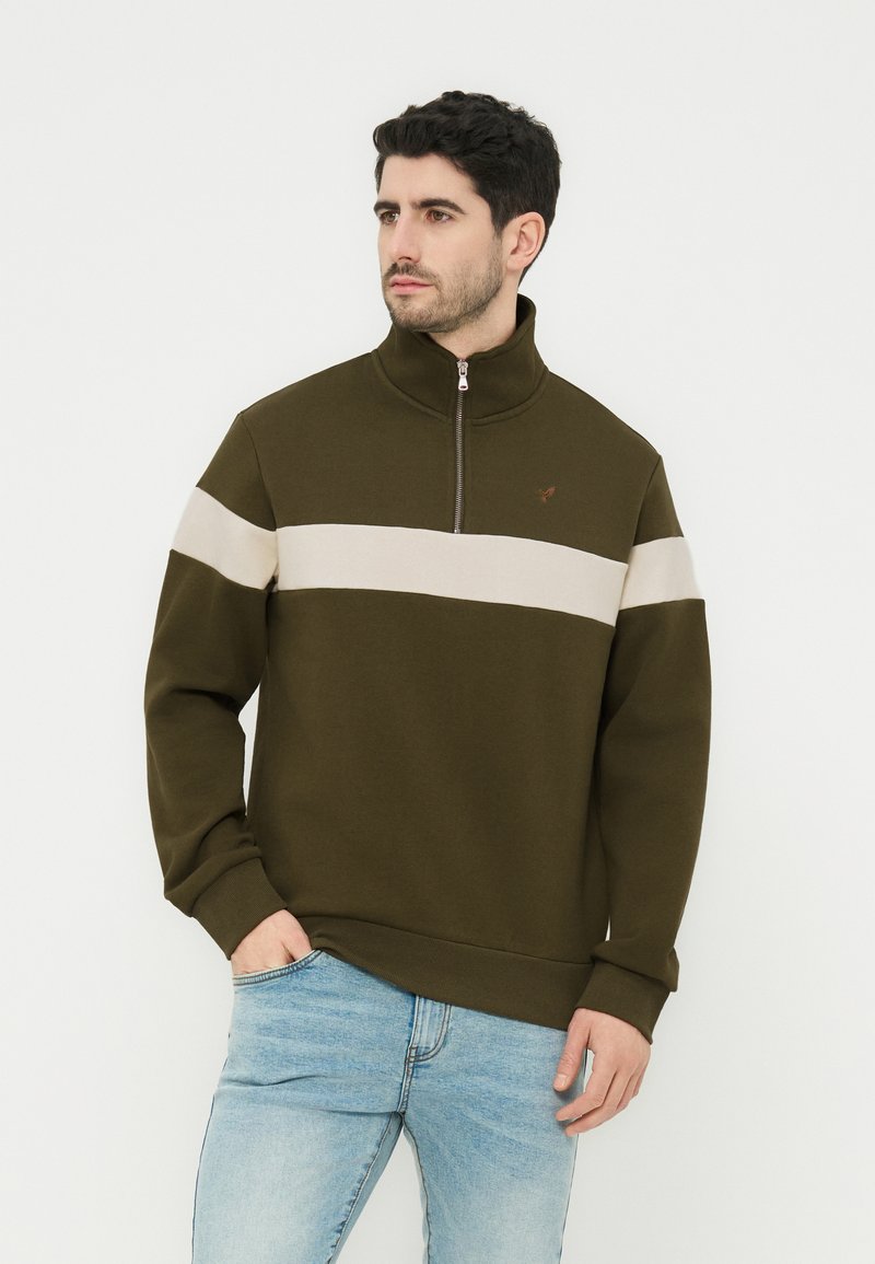 Άνδρας που φοράει ελιά πράσινο φούτερ με φερμουάρ μέχρι τη μέση και μπεζ οριζόντια ρίγα στο στήθος, συνδυασμένο με ανοιχτό μπλε τζιν, στέκεται μπροστά σε λευκό φόντο.