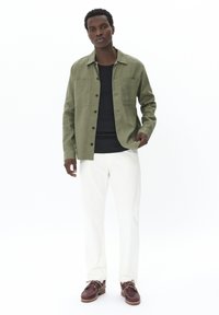 Camisa verde de botones con dos bolsillos frontales, camiseta negra por debajo, pantalones blancos y zapatos marrones. Textura suave, corte moderno y estilo casual.
