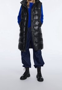 Schwarze Steppweste mit glänzender Oberfläche, Reißverschluss und Kapuze; kombiniert mit einem blauen Pullover, lockeren blauen Jeans und schwarzen Schnürstiefeln.