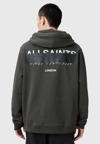 Donker olijf groene hoodie van katoenfleece, met een groot wit logo op de achterkant, met tekst in meerdere talen eronder.