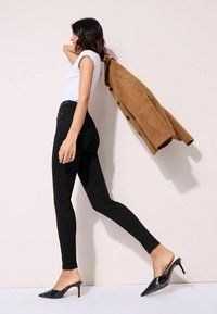 Fekete, slim-fit farmernadrág, fehér, testhez álló póló, bézs kabát az egyik karján, és fekete, magassarkú mules. Simító anyag és igényes kialakítás.
