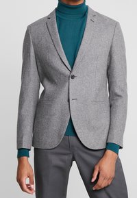 Man i grå blazer över en teal-färgad polotröja och grå byxor, stående mot en enkel ljus bakgrund.