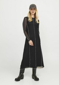 Zwarte maxi-jurk met lange doorschijnende mouwen, contrasterende witte stiksels en een vloeiende silhouet. Gemaakt met zwarte kniehoge laarzen en een pet.