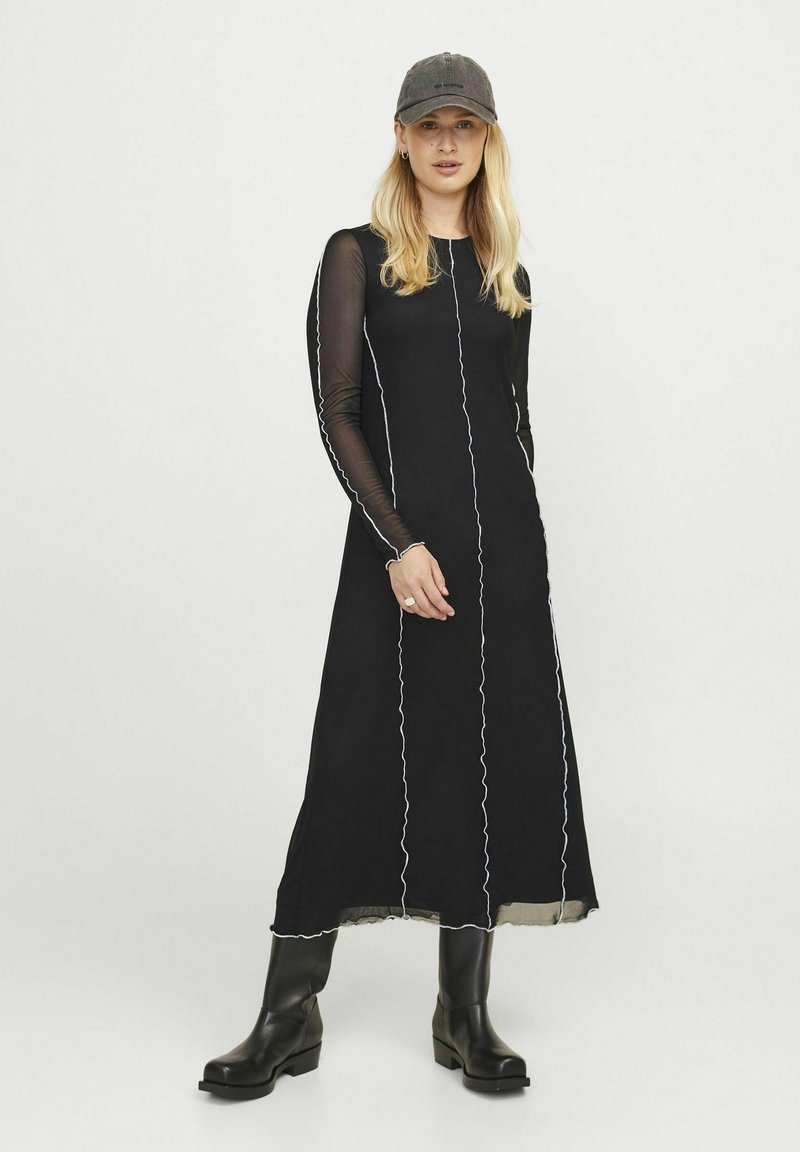 Zwarte maxi-jurk met lange doorschijnende mouwen, contrasterende witte stiksels en een vloeiende silhouet. Gemaakt met zwarte kniehoge laarzen en een pet.
