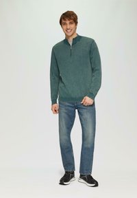 Heren zip-nek trui in gemêleerd teal stof, lange mouwen, geribbelde manchetten en onderrand, gecombineerd met lichtblauwe jeans en zwarte sneakers.