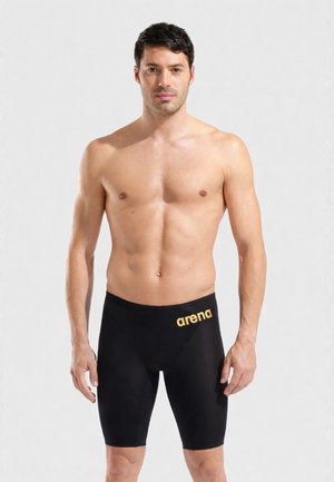 Hombre sin camiseta vistiendo jammers de natación negros de Arena con logo dorado, de pie contra un fondo claro y liso, mirando hacia adelante.