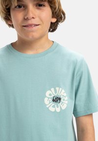 Camiseta de algodón azul claro con un logo circular que incluye el texto "SQUIRREL SILVER" y un gráfico estilizado de un animal. Manga corta, cuello redondo.
