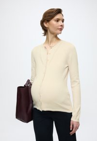 Cream cardigan with buttons, soft texture, long sleeves, layered over a fitted black top. Dark burgundy handbag with a smooth finish.