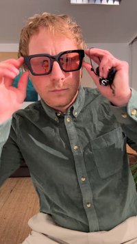 Óculos de sol oversized pretos com uma armação quadrada e lentes laranjas. O usuário segura as hastes e está vestido com uma camisa de veludo cotelê verde.