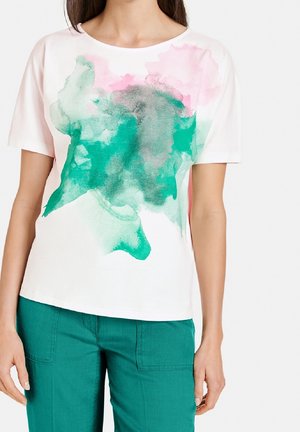 Femme portant un t-shirt blanc à manches courtes avec un imprimé aquarelle vert et rose, associé à un pantalon sarcelle sur un fond uni.