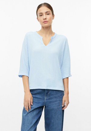 Frau mit zurückgebundenen Haaren, die ein hellblaues, strukturiertes Oberteil mit Dreiviertelärmeln und blaue Jeans trägt, steht vor einem schlichten weißen Hintergrund.