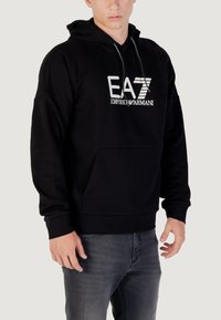 Felpa nera in cotone con tasca a marsupio anteriore, cappuccio con cordino e logo EA7 Emporio Armani in bianco. Vestibilità standard con polsini a costine.