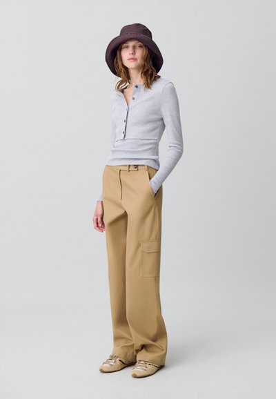 Maglione grigio a maniche lunghe con dettagli a bottone, abbinato a pantaloni larghi beige con tasche laterali e un cappello a secchiello marrone. Calzature casual visibili.