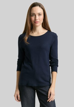 Femme aux longs cheveux bruns portant un haut à manches longues bleu marine et un jean foncé, debout devant un fond gris uni.