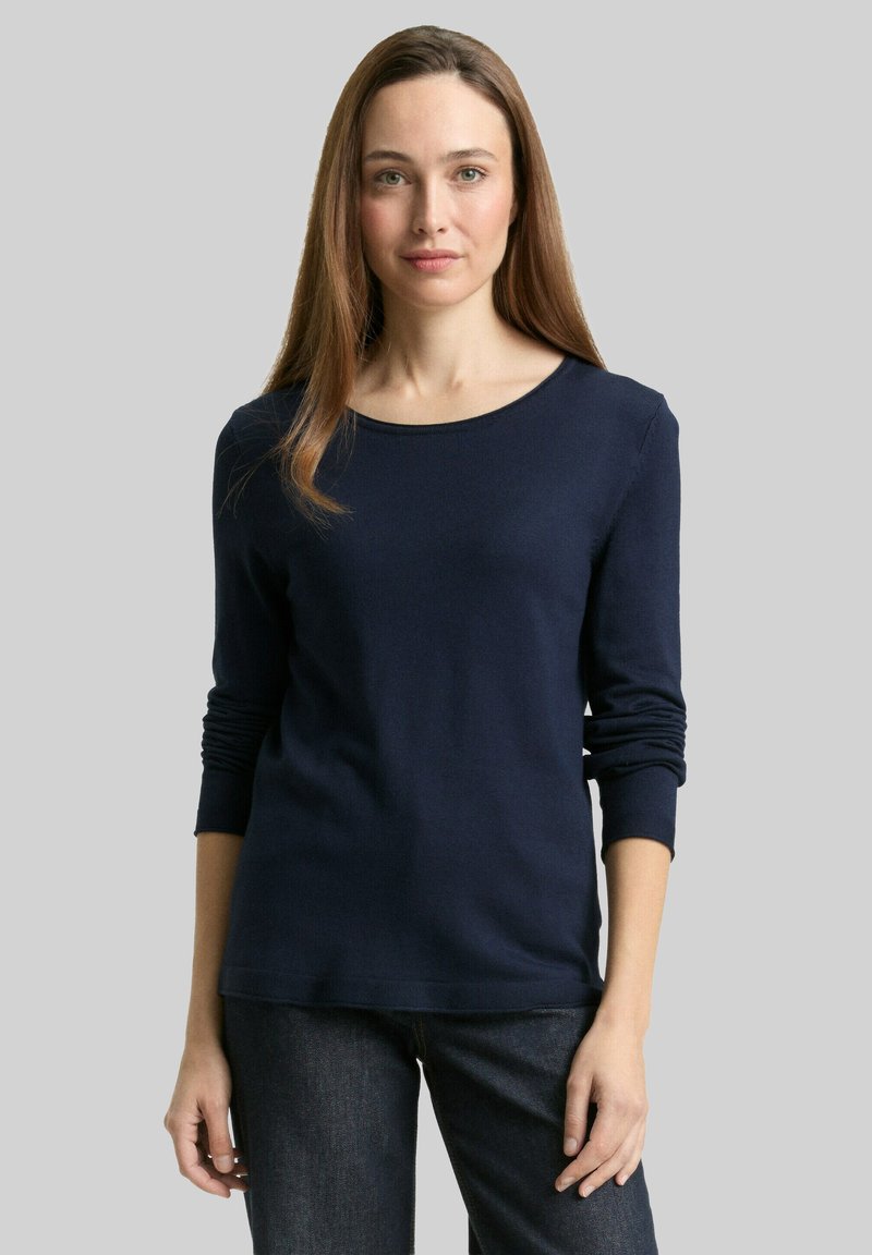 Femme aux longs cheveux bruns portant un haut à manches longues bleu marine et un jean foncé, debout devant un fond gris uni.