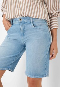 Shorts en denim bleu clair avec une coupe droite, cinq poches et une fermeture à bouton, associés à une chemise à manches courtes à rayures beiges et blanches.