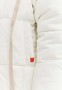 Biała kurtka puchowa z pionowymi przeszyciami, posiadająca beżowy zamek błyskawiczny i małą czerwoną metkę z logo na kieszeni. Miękki, teksturowany materiał.