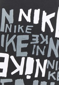 Černé bavlněné tričko s dynamickou typografií, na němž je slovo "NIKE" v bílé a šedé barvě, překrývající se v různých velikostech a orientacích.