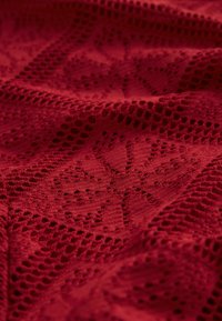 Tissu tricoté rouge avec un motif en treillis ouvert comportant des motifs floraux. La texture semble douce, avec des détails intriqués et une profondeur de couleur riche.
