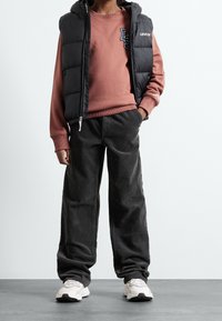 Schwarze Steppweste über einem staubroten Sweatshirt mit Logo, kombiniert mit weiten schwarzen Cordhosen und hellen Turnschuhen.