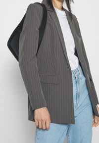 Persona che indossa un blazer grigio a righe, una t-shirt bianca e jeans azzurri chiari, con una borsa a tracolla nera sul lato sinistro.