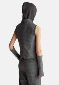 Szary w kratę skrócony kamizelka z wysokim kołnierzem i przymocowaną ciemnoszara dzianinową kapturem. Długie rękawy z prążkowanego materiału w dopasowanym szarym kolorze. Dopasowany krój.