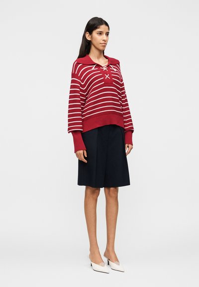 Γυναίκα που στέκεται φορώντας μακρυμάνικη μπλούζα με κόκκινες και λευκές ρίγες, μαύρη φούστα μήκους μέχρι το γόνατο και λευκά μυτερά τακούνια σε απλό φόντο.