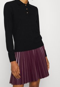 Polo de manga larga negro con cierre de botones, combinado con una falda plisada de cuero burdeos de longitud a la rodilla. Detalle de material texturizado.