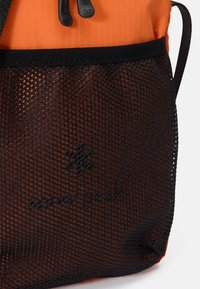 Zaino arancione con una tasca in rete nera, spallacci regolabili e chiusura con cordino. Materiali leggeri e testurizzati aumentano la durata.