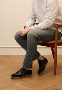 Sneaker nera con tomaia in suede e pelle, suola in gomma e lacci frontali. Indossata con jeans blu chiaro e calzini bianchi, seduta su una sedia di legno.