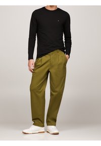 Schwarzes Langarmshirt und olivgrüne, plissierte Hose. Weiße Sneaker mit Gummisohlen. Glatter Stoff, lockerer Schnitt und maßgeschneiderte Passform.