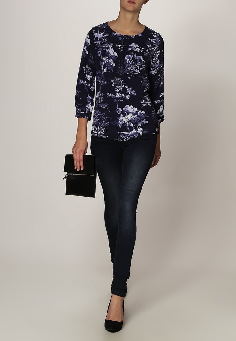 Blusa floreale blu navy con maniche a tre quarti, abbinata a jeans skinny scuri e una clutch nera. Presenta intricati motivi floreali.